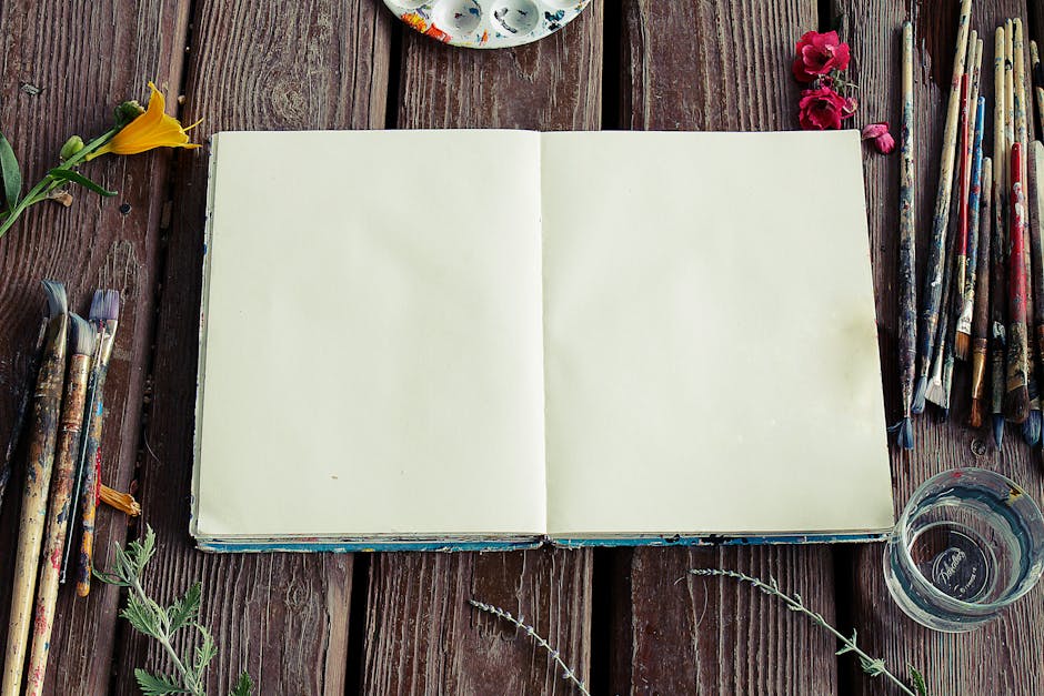 Blank sketchbook surrounded by paintbrushes and flowers on rustic wooden surface.