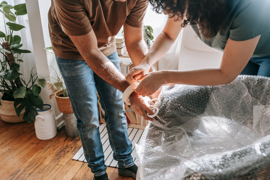 A couple wraps furniture with bubble wrap in a sunlit living room, preparing to move.
